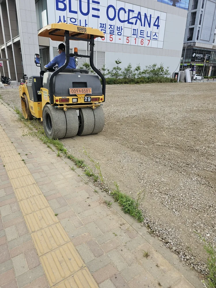 노란색 로드롤러가 보도 옆 골재 바닥을 눌러 다지는 주차장 공사 현장