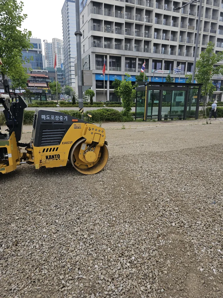 노란색 로드롤러가 골재가 깔린 주차장 부지를 다지는 모습