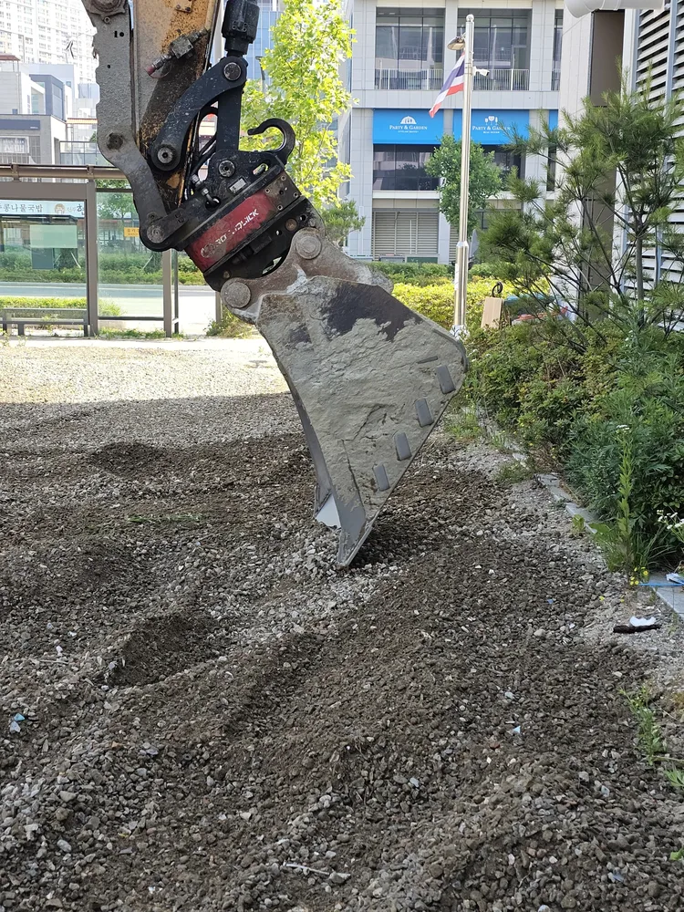 굴착기 버킷이 흙과 자갈이 섞인 부지 표면을 걷어내는 주차장 부지 정리 작업